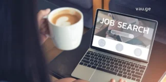 სტუდენტობიდან კარიერულ ჩიხამდე, როგორ იცვლება დასაქმების პარადიგმა საქართველოში A young person holding a coffee cup and searching for jobs on a laptop with "Job Search" on the screen.