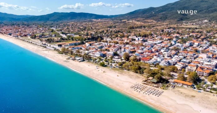 Aerial view of a wide sandy beach and turquoise sea in Halkidiki, Greece, with coastal town and mountains.
