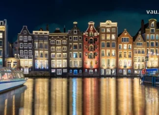 ამსტერდამი: ევროპული ვოიაჟი არხებისა და თავისუფლების სამყაროში Illuminated historic canal houses in Amsterdam at night with golden reflections on the water and a glass-top tour boat.