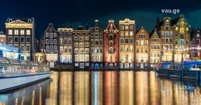 Illuminated historic canal houses in Amsterdam at night with golden reflections on the water and a glass-top tour boat.
