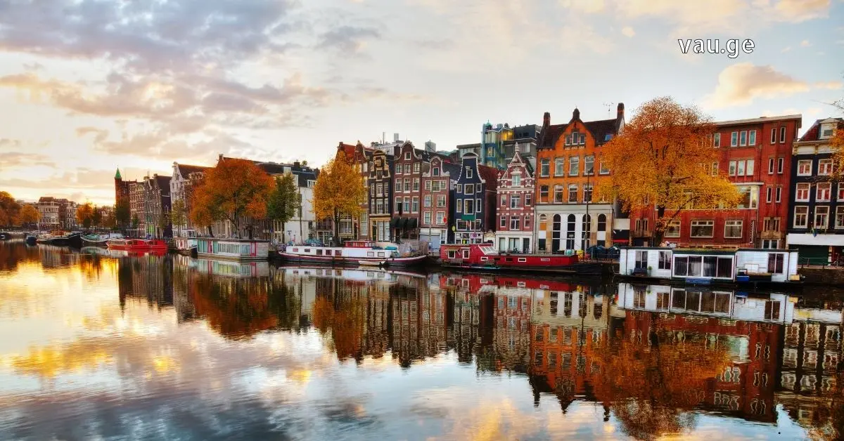 Autumn sunset in Amsterdam featuring colorful canal houses and houseboats reflected in the calm water under a cloudy sky.