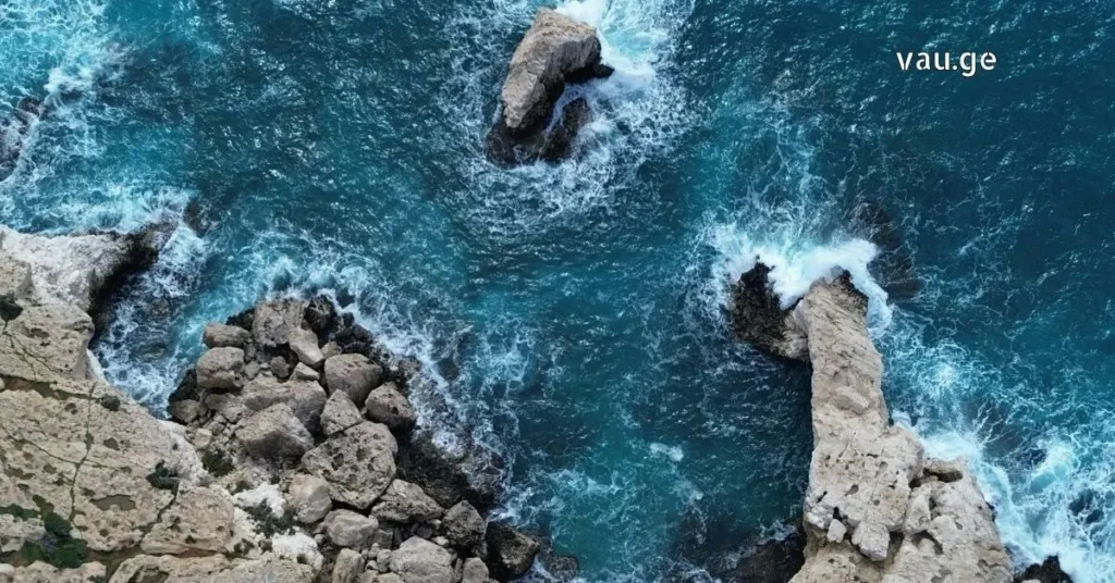 Top-down aerial view of a rugged, light-tan coastline with sea stacks and crashing turquoise waves.
