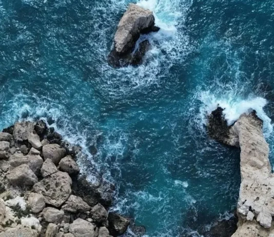 აია ნაპა: ხმელთაშუა ზღვის მარგალიტი – თქვენი დაუვიწყარი საზაფხულო თავგადასავალი Top-down aerial view of a rugged, light-tan coastline with sea stacks and crashing turquoise waves.